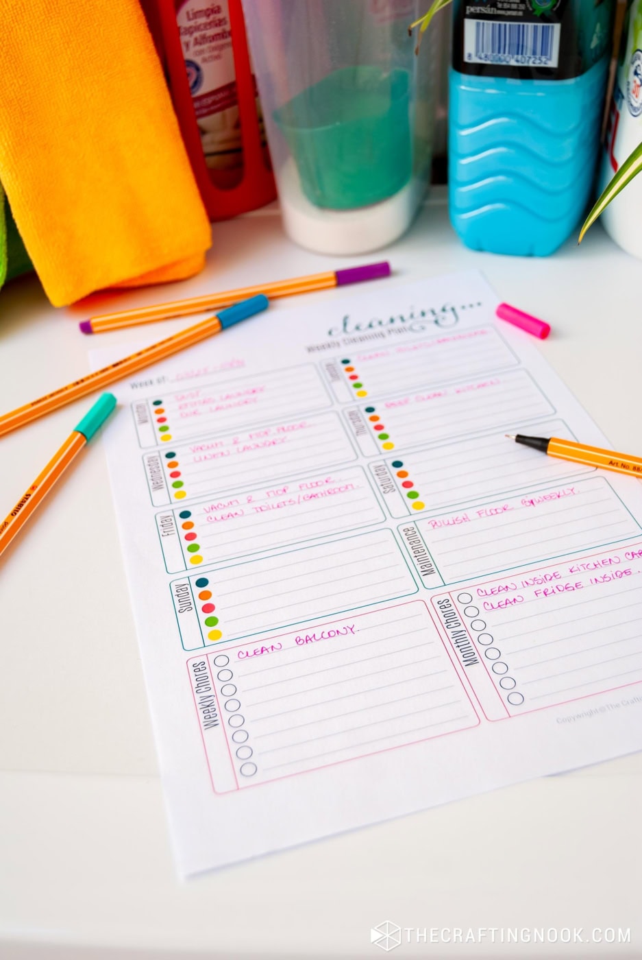 45 degree angle view of Fun Free Printable Weekly Cleaning Schedule sheet laying on top of washer machine with markers around and written tasks on it with focus point on weekly and monthly tasks