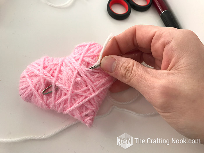 Inserting a yarn string to the back of a pink heart to attach ir to the wreath