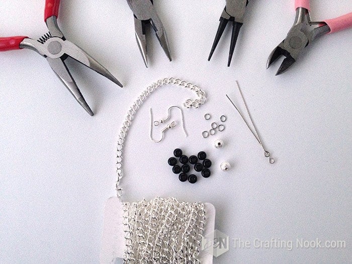 Jewelry-making supplies for DIY triangle dangle earrings, including black and silver beads, chain, eye pins, earring hooks, jump rings, and pliers.
