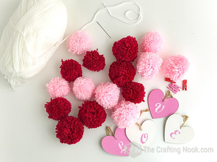 White yarn, lots of pink and red pom poms, wooden heart ornaments and clothespins. Supplies to make this Pom Pom Garland.