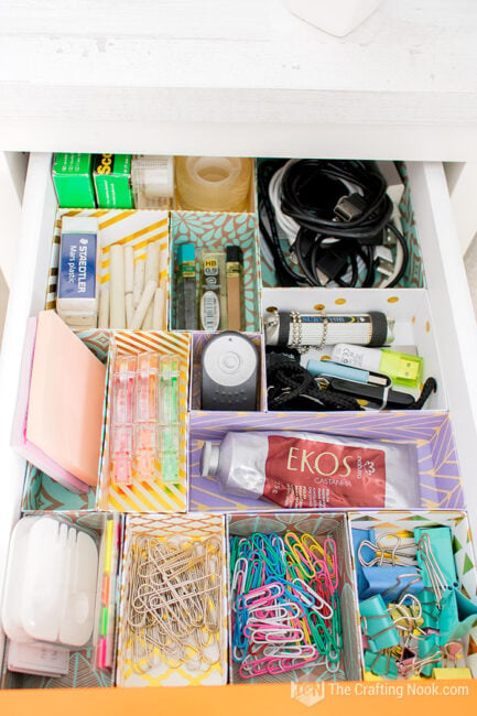 Open desk drawer organized with cardstock compartments for cables, small supplies, stationery, and office tools.