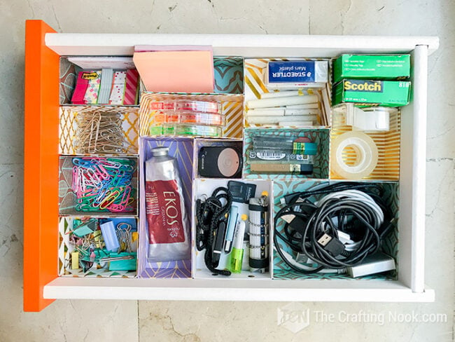 Organized desk drawer filled with custom patterned cardstock compartments holding office supplies, cables, tape, and small tools.