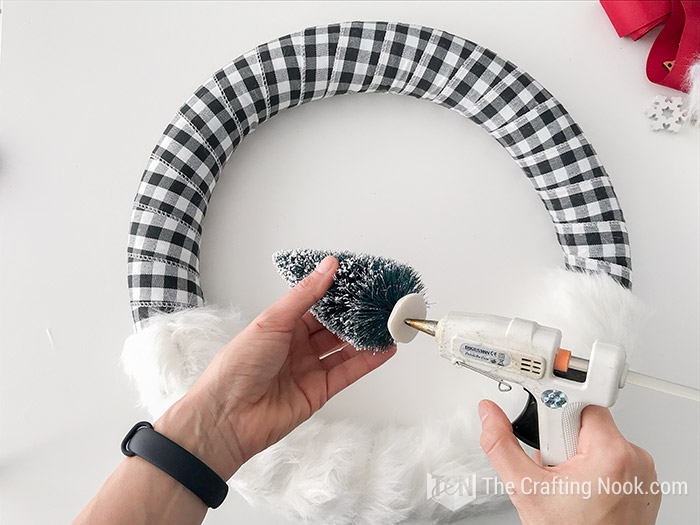 Applying hot glue to the base of a pine tree to attach it to the wreath