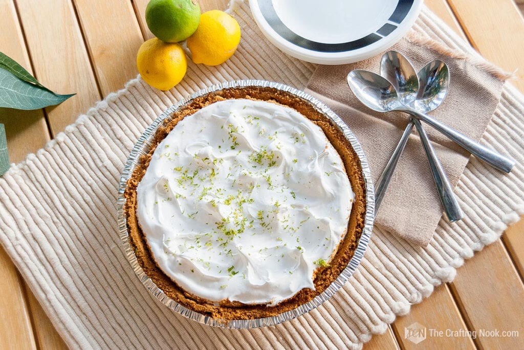 A key lime pie topped with whipped cream and lime zest is placed on a beige woven mat on a wooden table. Nearby are three fresh limes, two white bowls, and three spoons on a beige napkin. Green leaves are in the corner, adding a touch of freshness.