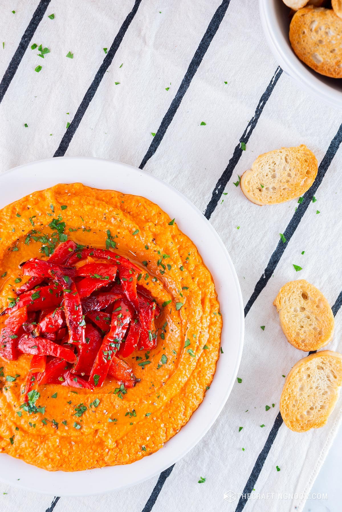 Homemade Roasted Red Pepper Hummus Recipe in a dish decorated with roasted pepper strips on the table with tosts around the dish