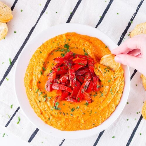 Homemade Roasted Red Pepper Hummus Recipe in a dish decorated with roasted pepper strips and me dipping a toast. Feature image