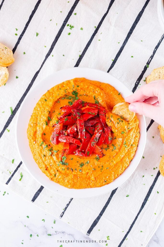 Homemade Roasted Red Pepper Hummus Recipe in a dish decorated with roasted pepper strips and me dipping a toast. Feature image