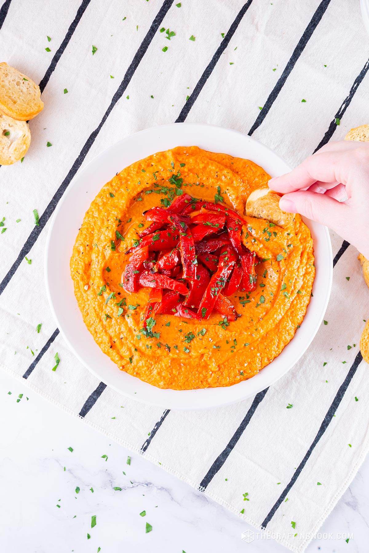 Homemade Roasted Red Pepper Hummus Recipe in a dish decorated with roasted pepper strips and me dipping a toast