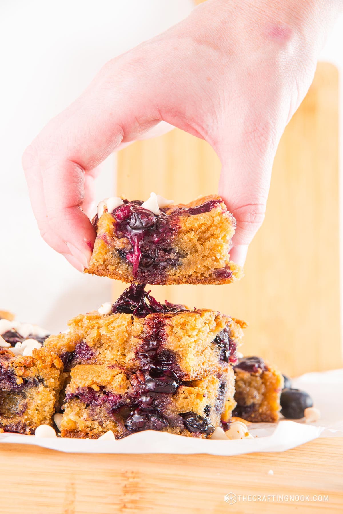 side view of a hand grabbing a piece White Chocolate Blueberry Blondies cut in nineths on a piece ofpartchment paper and a cutting board.