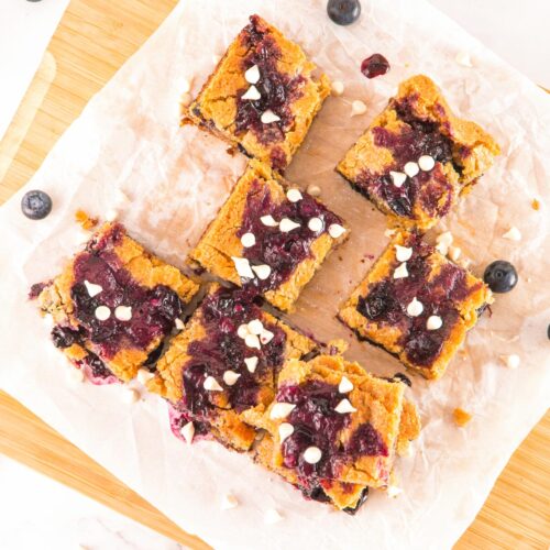 Scattered pieces of White Chocolate Blueberry Blondies cut in nineths on a piece ofpartchment paper and a cutting board. Cover image