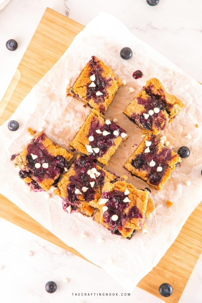 Scattered pieces of White Chocolate Blueberry Blondies cut in nineths on a piece ofpartchment paper and a cutting board. Cover image