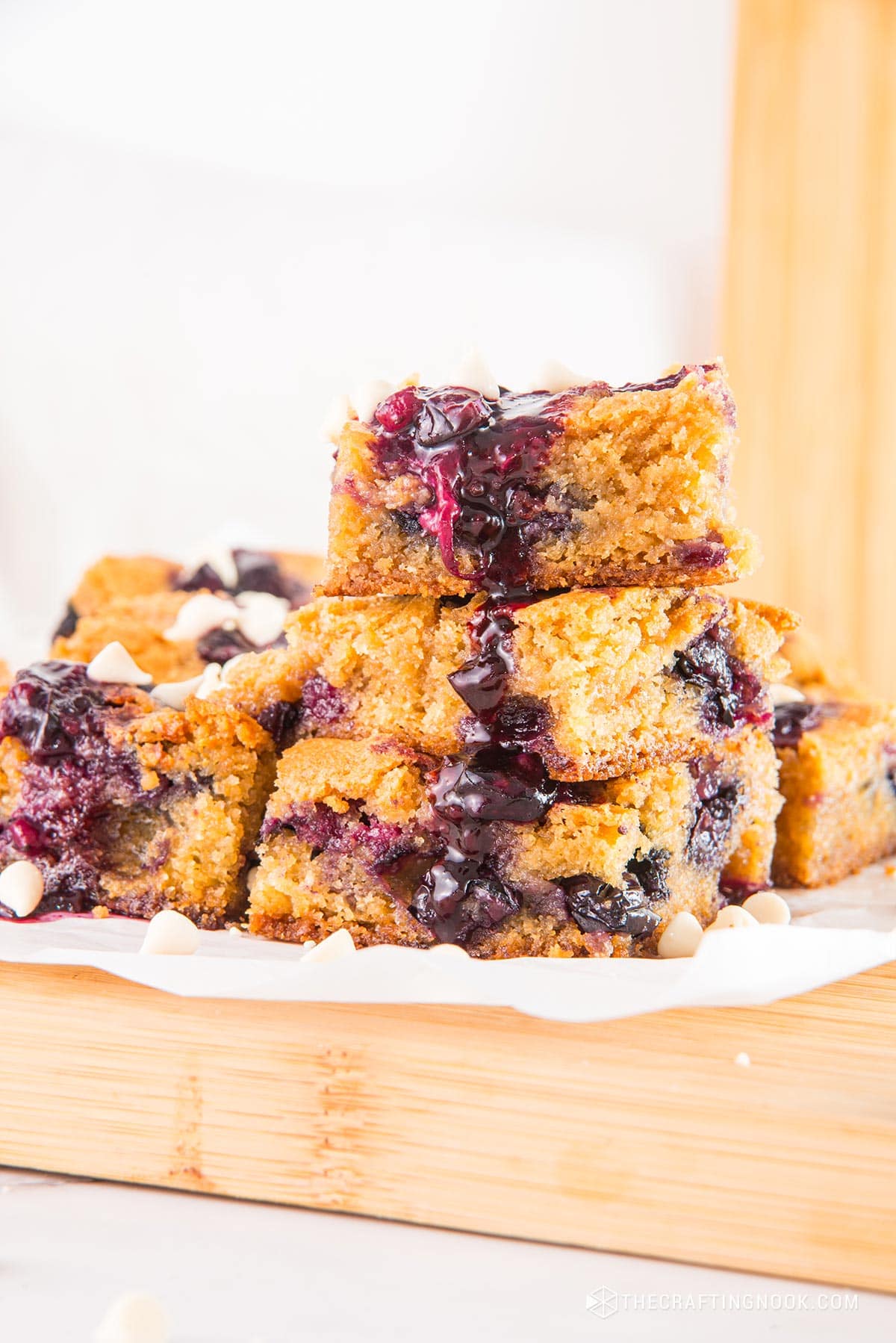 Stacked pieces of White Chocolate Blueberry Blondies cut in nineths on a piece ofpartchment paper and a cutting board.