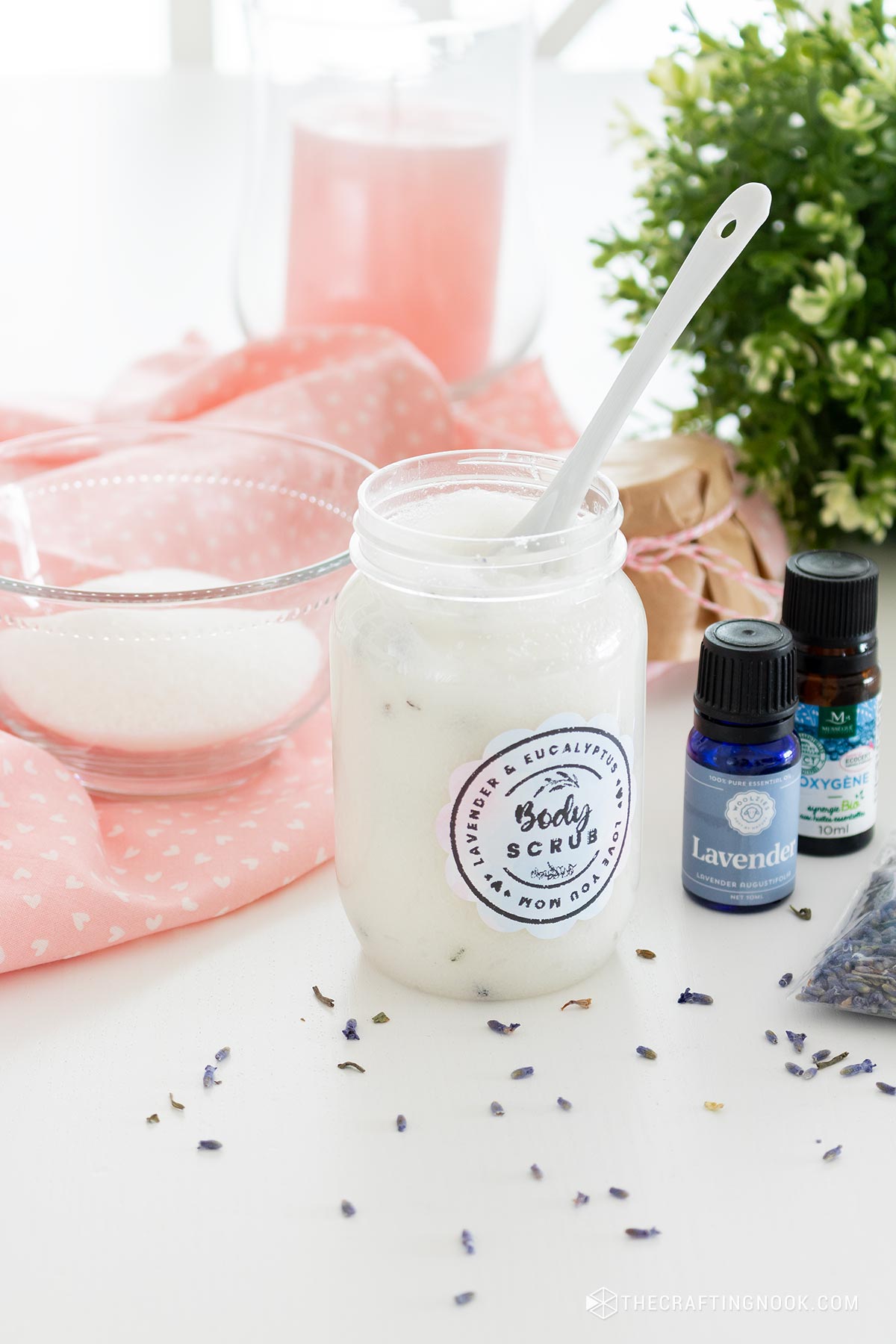 Eucalyptus Lavender Sugar Scrub Recipe in a jar with a spoon inside surrounded but props like a tawel, suggar and a plant