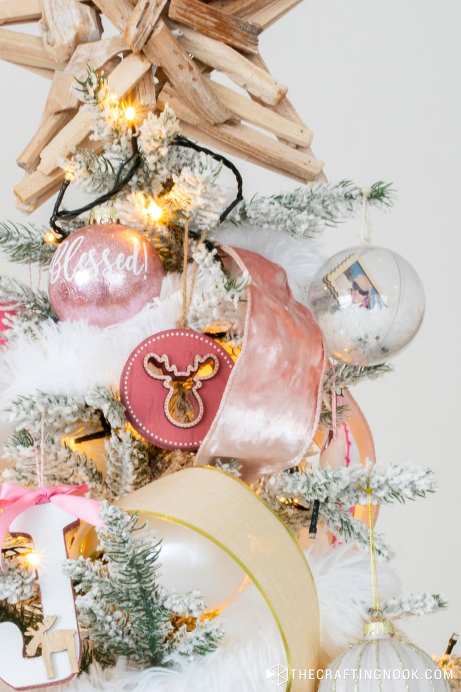 Close to the top of the tree with snow gllobe ornament, a pink glitter ornament written Blessed on it and a pink wooden ornemante with a deer cutout