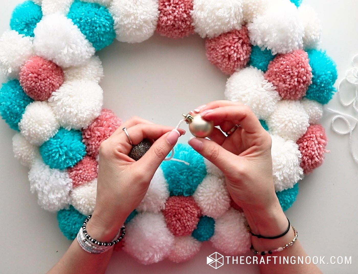 close up image of hands holding a gold ornaments to place them on the pompom garland
