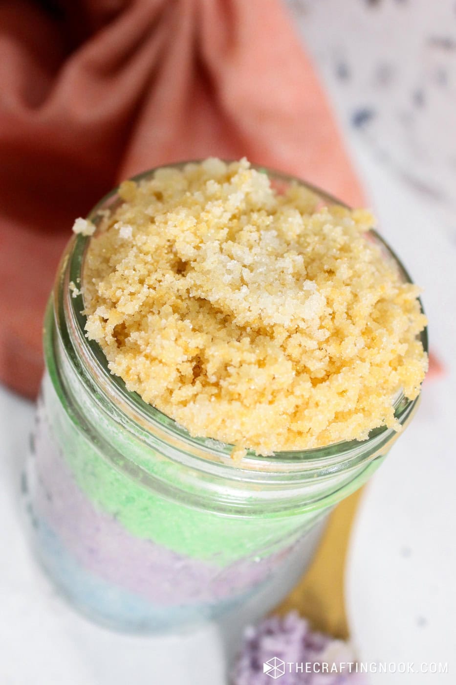 Closeup in an 45 degre angle of the jar layers of sugar scrub in different pastel colors, blue, purple, green and yellow.