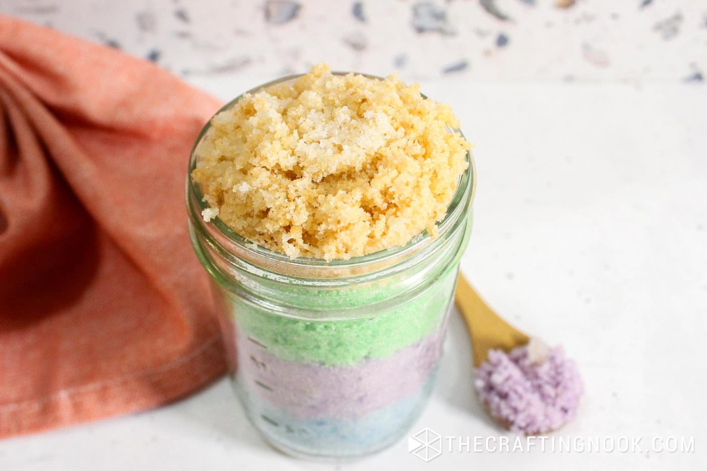 A 45 degre angle view of the jar layers of sugar scrub in different pastel colors, blue, purple, green and yellow.