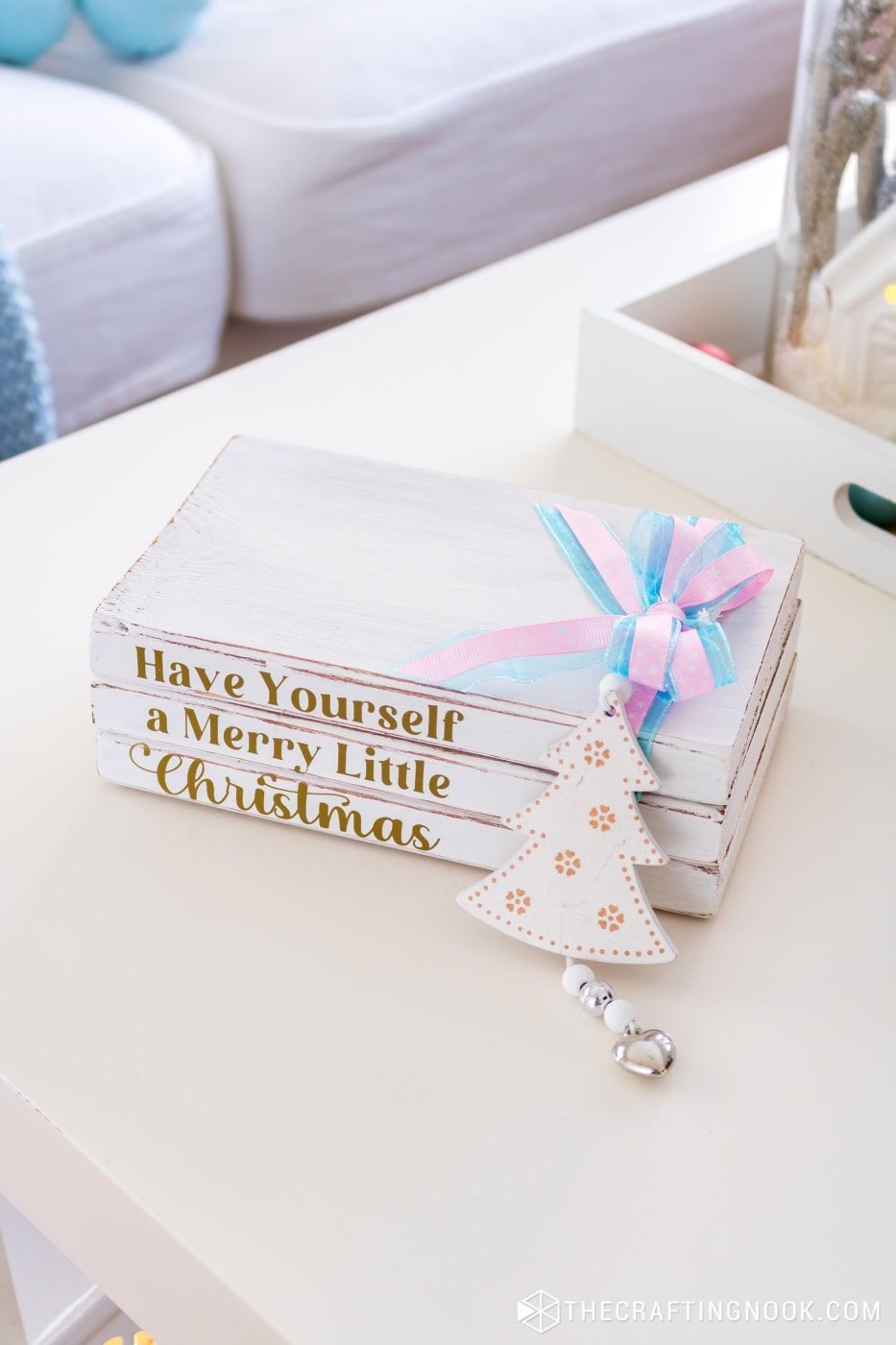 Wooden Christmas book stack with gold vinyl lettering and a pastel ribbon bow displayed on a white table with a soft cozy background.