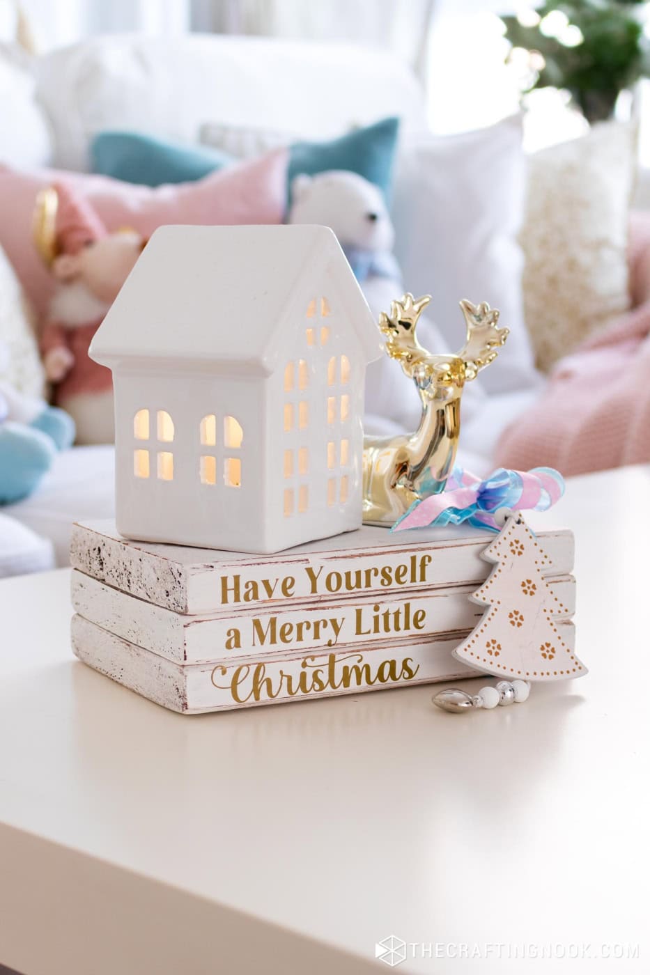 Christmas wooden book stack decorated with a bow and tree ornament, displayed with a white ceramic house and gold reindeer.