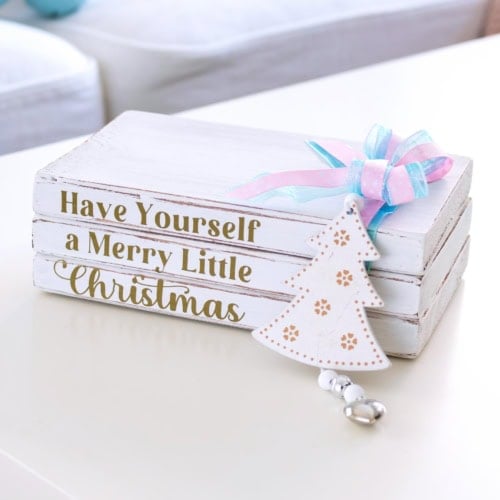 DIY Christmas wooden book stack with gold lettering and pastel ribbon sitting on a white table in a cozy living room. Feature Image.