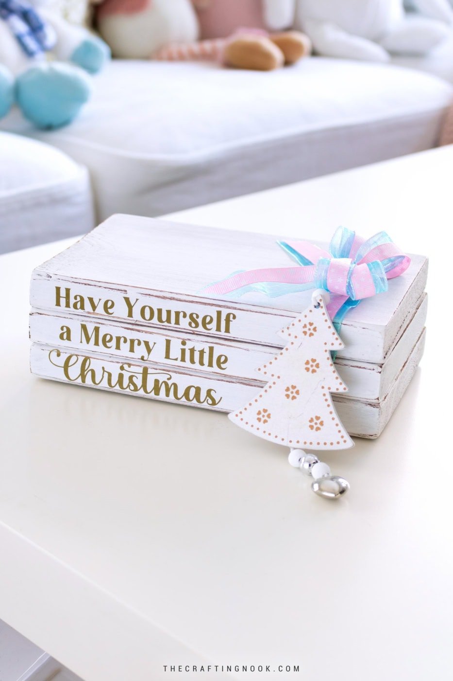 DIY Christmas wooden book stack with gold lettering and pastel ribbon sitting on a white table in a cozy living room. Feature Image.