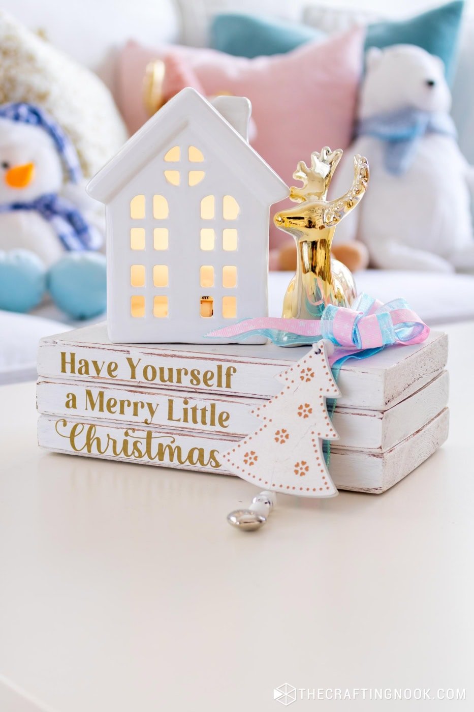 Close-up of the wooden book stack with gold vinyl Christmas text, white ceramic house, and gold reindeer dรฉcor in a pastel living room.