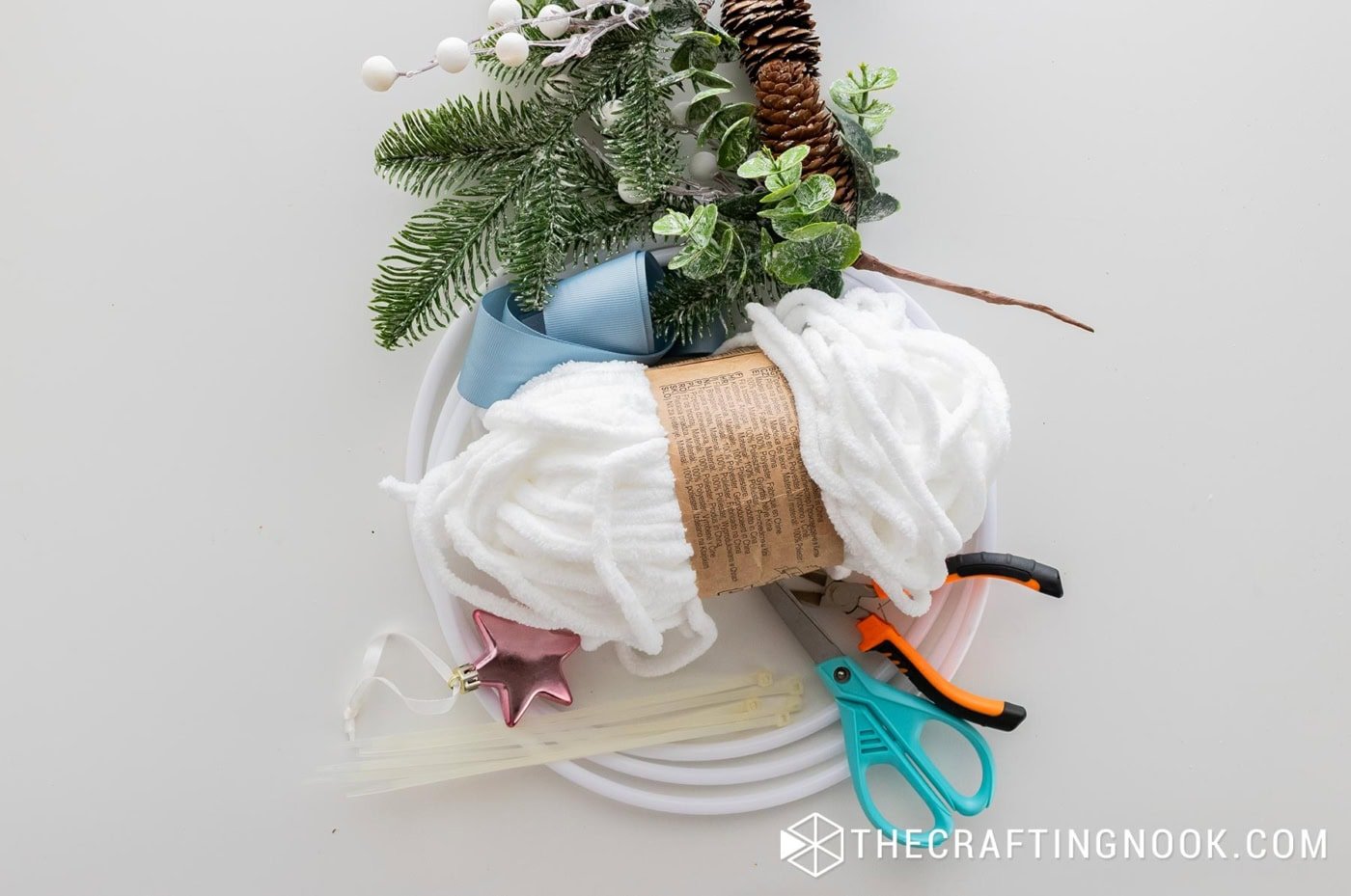 Supplies laid out on a table including three white hoops, white chenille yarn, faux pine greenery, pinecones, berries, blue ribbon, a small pink star ornament, zip ties, scissors, and wire cutters.