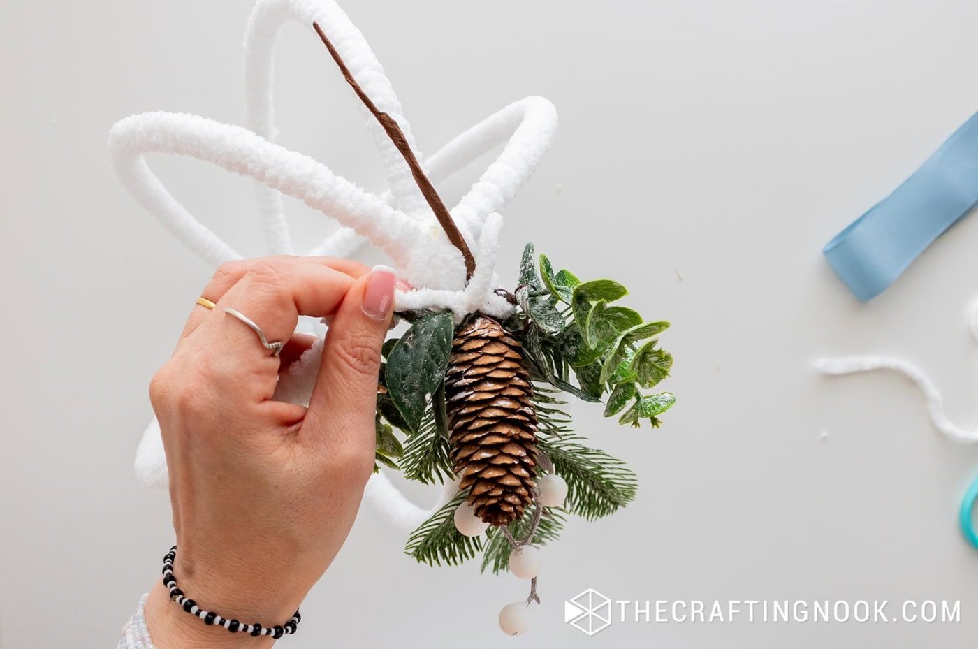 Attaching faux greenery and a pinecone to the yarn-wrapped hoop using yarn instead of glue.
