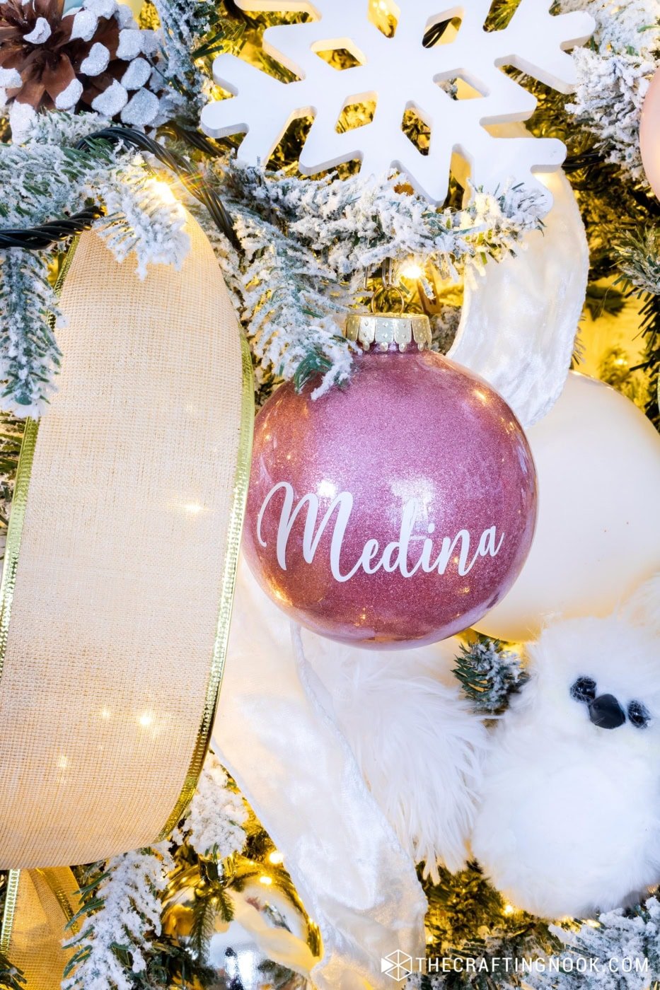 Handmade glitter ornament with a name on vinyl placed among flocked branches and soft ribbon.