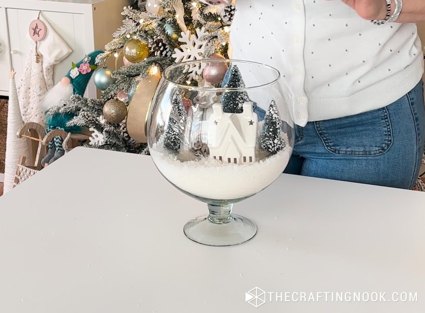 Bottle brush trees being added behind the ceramic house inside the glass vase. A small metallic deer figurine being set next to the house inside the snowy vase scene.