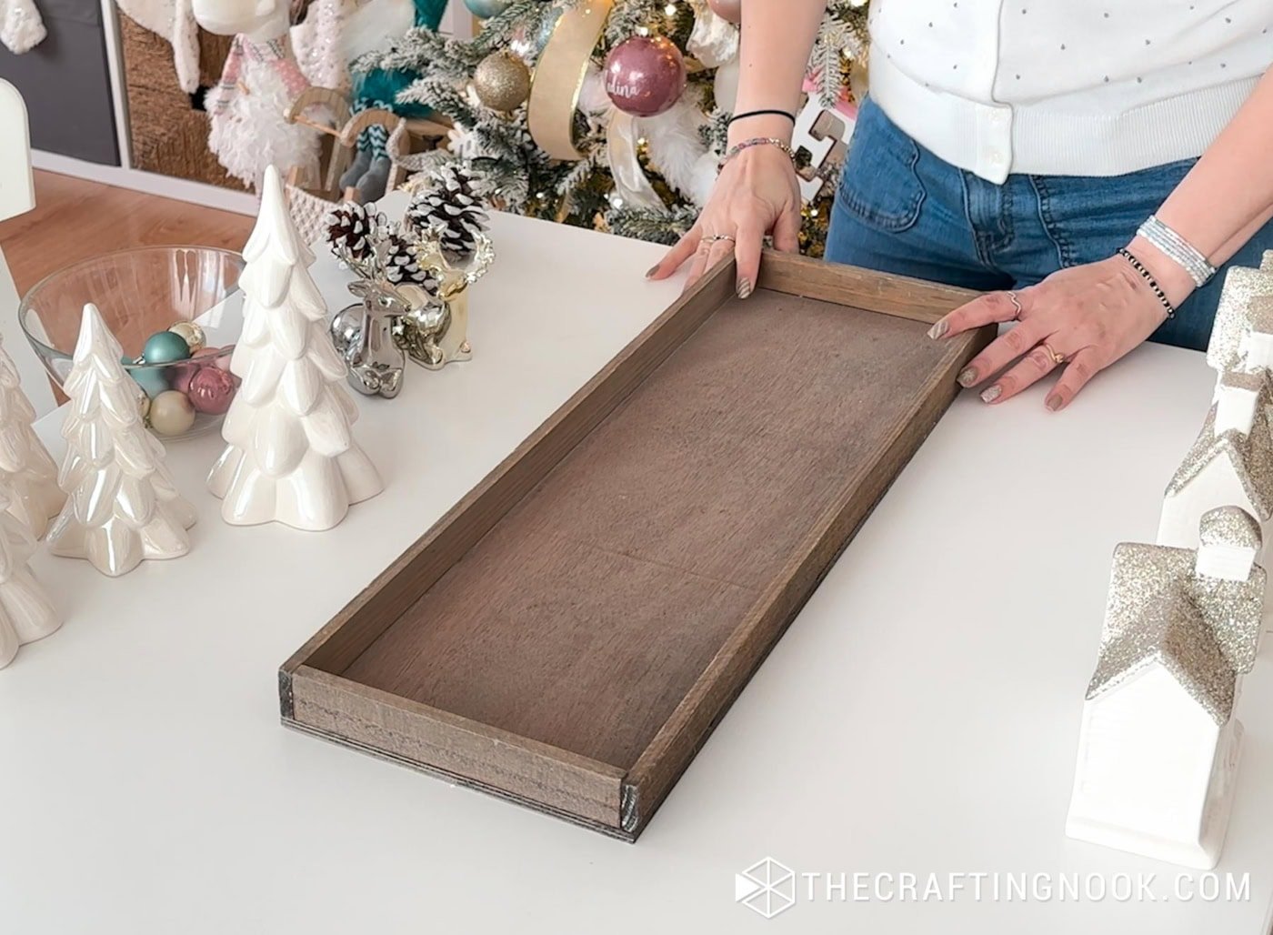 Hands placing an empty wooden tray on a white table, surrounded by ceramic trees and Christmas decorations.