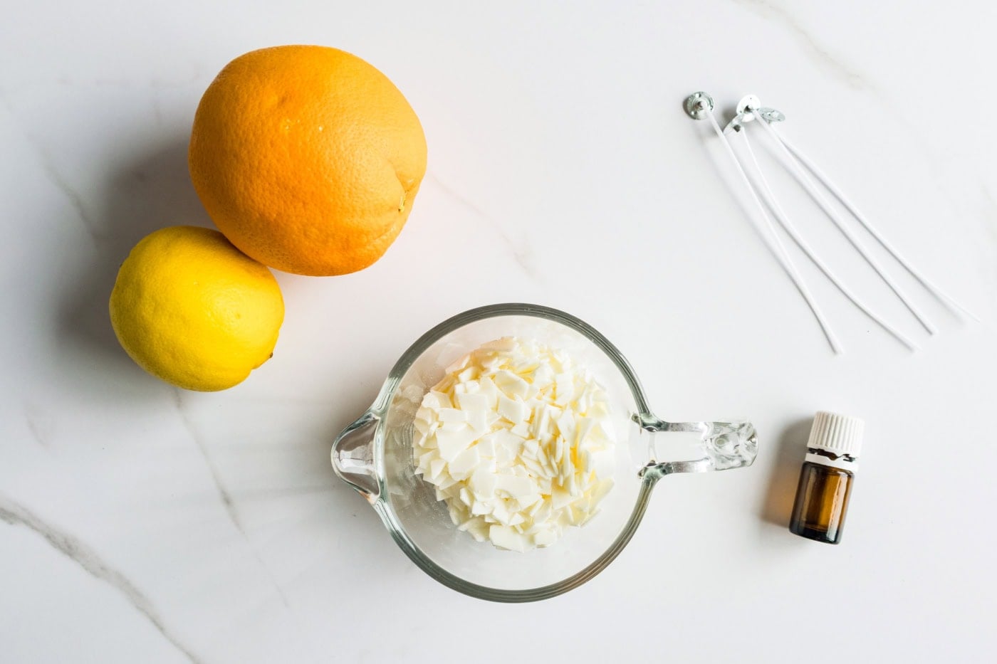 Supplies for DIY citrus peel candles, including an orange, lemon, soy wax flakes, pre-tabbed wicks, and fragrance oil.