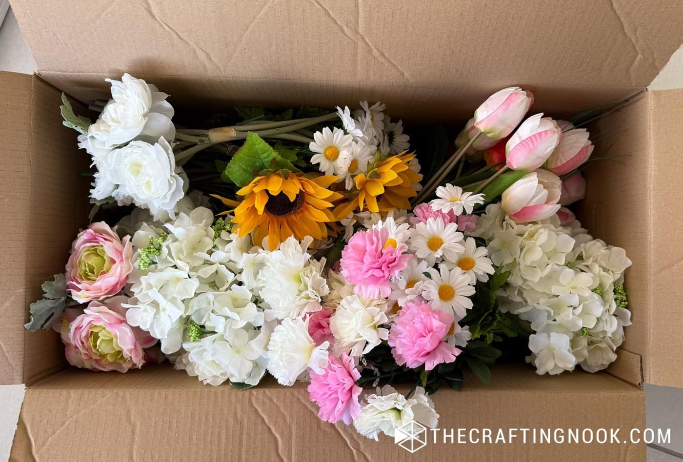Cardboard box filled with faux flowers including sunflowers, hydrangeas, daisies, and pink tulips