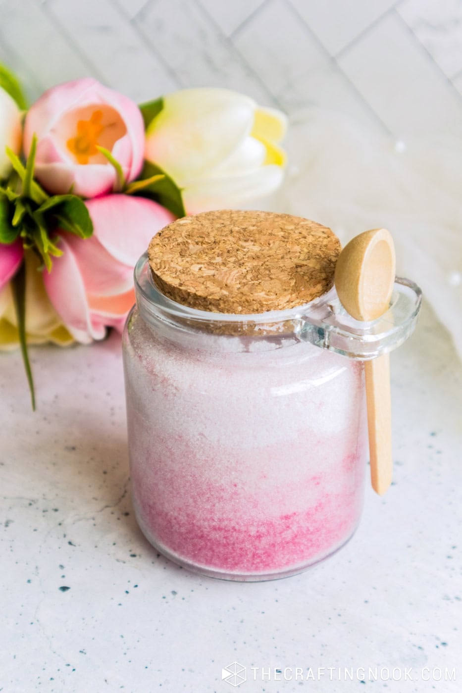 Cork lid jar of pink ombre sugar scrub with a wooden spoon, flowers and a succulent in the background