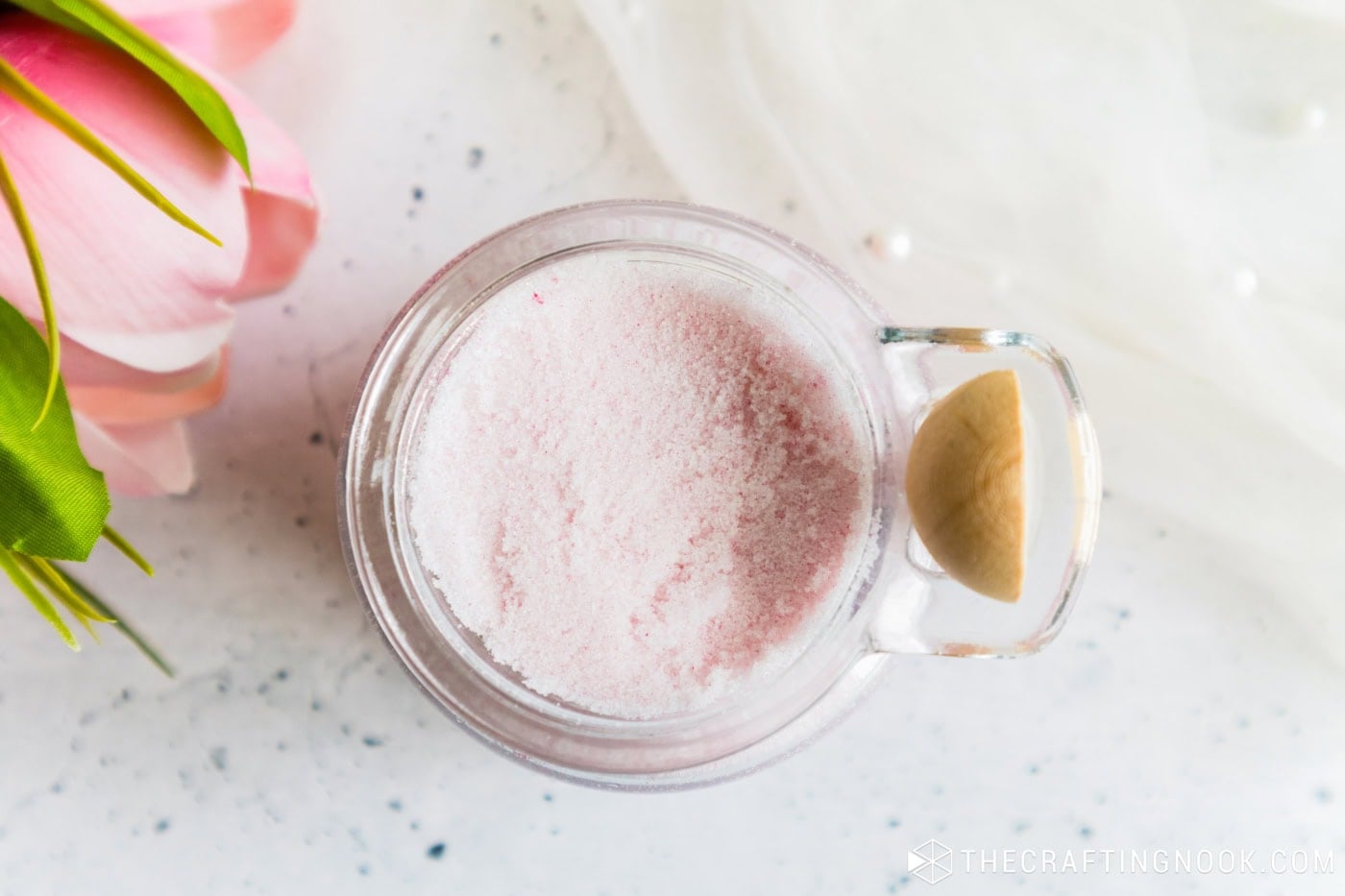 Top view of a jar of light pink sugar scrub with a wooden spoon on the handle and a pink tulip beside it