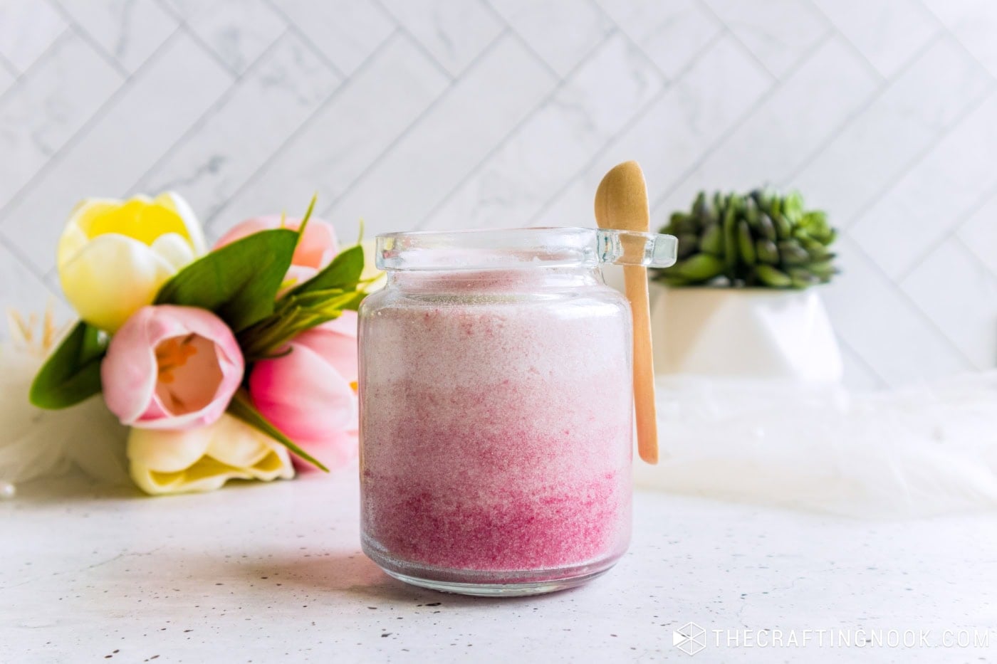 Open glass jar of pink ombre sugar scrub with a mini wooden spoon, tulips and a succulent