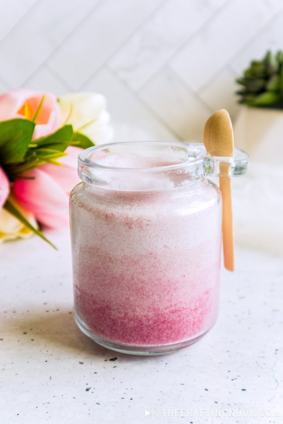 Open jar of pink ombre sugar scrub, seen from a side angle, with a wooden spoon hanging on the rim