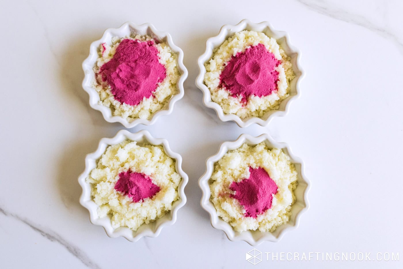 Four small white bowls of sugar scrub base, each topped with a different amount of pink powder