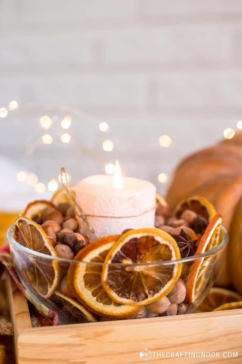 White lit candle in a glass bowl surrounded by dried orange slices, hazelnuts, and star anise