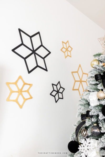 Five handmade black and gold popsicle stick snowflakes in different sizes arranged on a white wall.