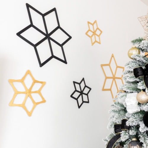 Five handmade black and gold popsicle stick snowflakes in different sizes arranged on a white wall.