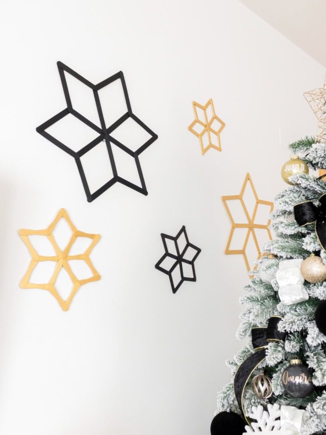 Five handmade black and gold popsicle stick snowflakes in different sizes arranged on a white wall.