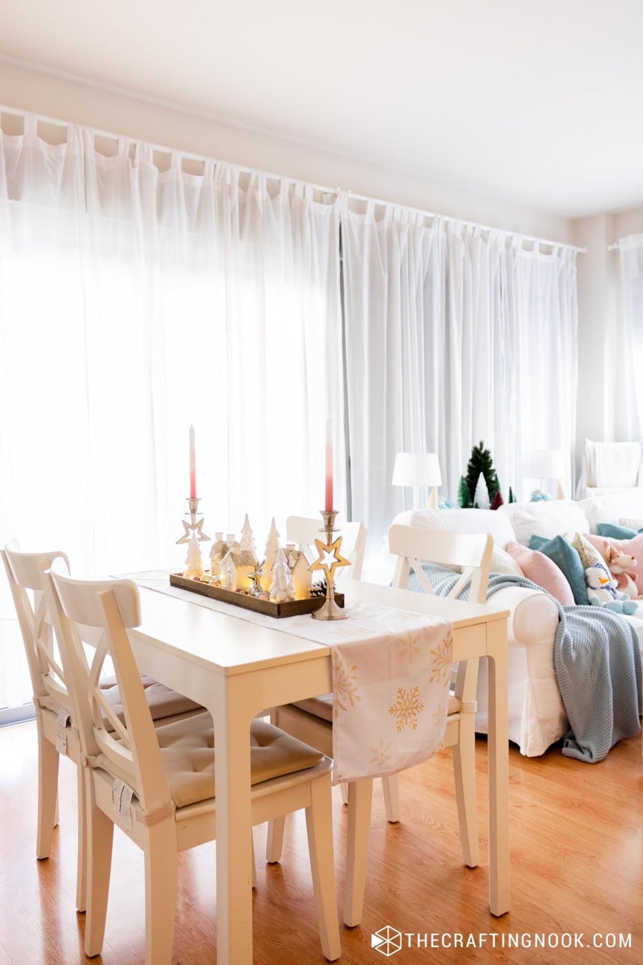 White dining table with a snowflake runner and a Christmas village centerpiece with pink candles, and a pastel Christmas sofa in the background