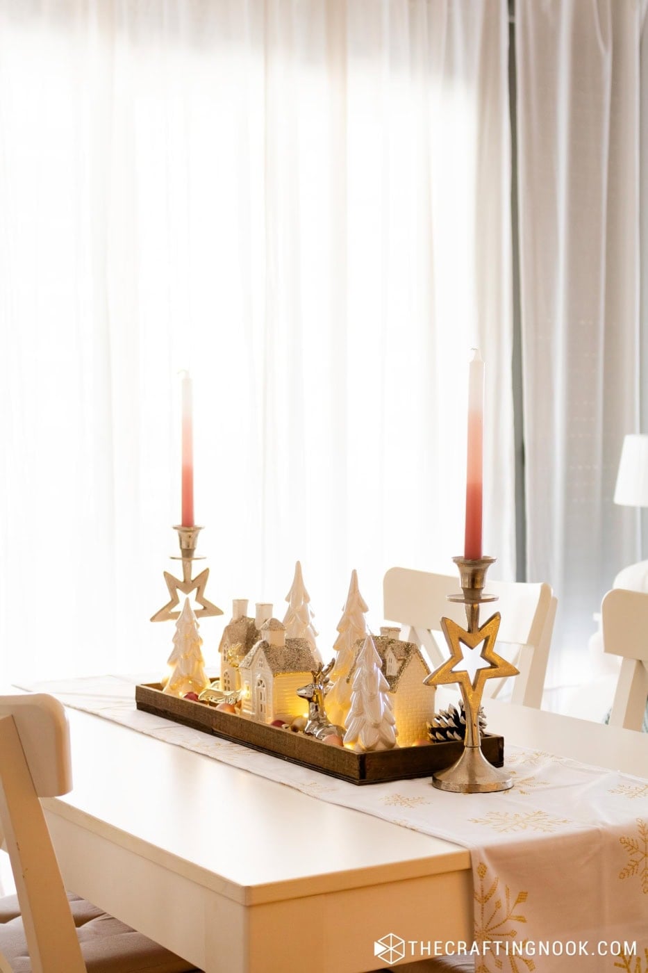 Wooden tray with white glitter houses, pearly trees and a silver reindeer between two silver star candle holders with pink candles