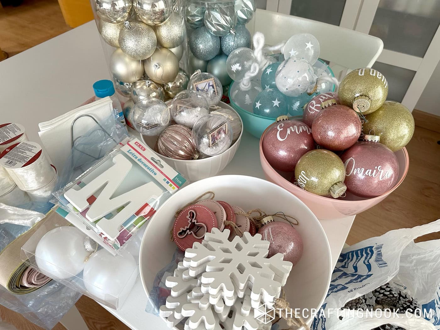White table with bowls of Christmas ornaments: glitter balls with white names, clear fillable balls with photos and snow, and wooden snowflakes, plus a wooden letter M and ribbon