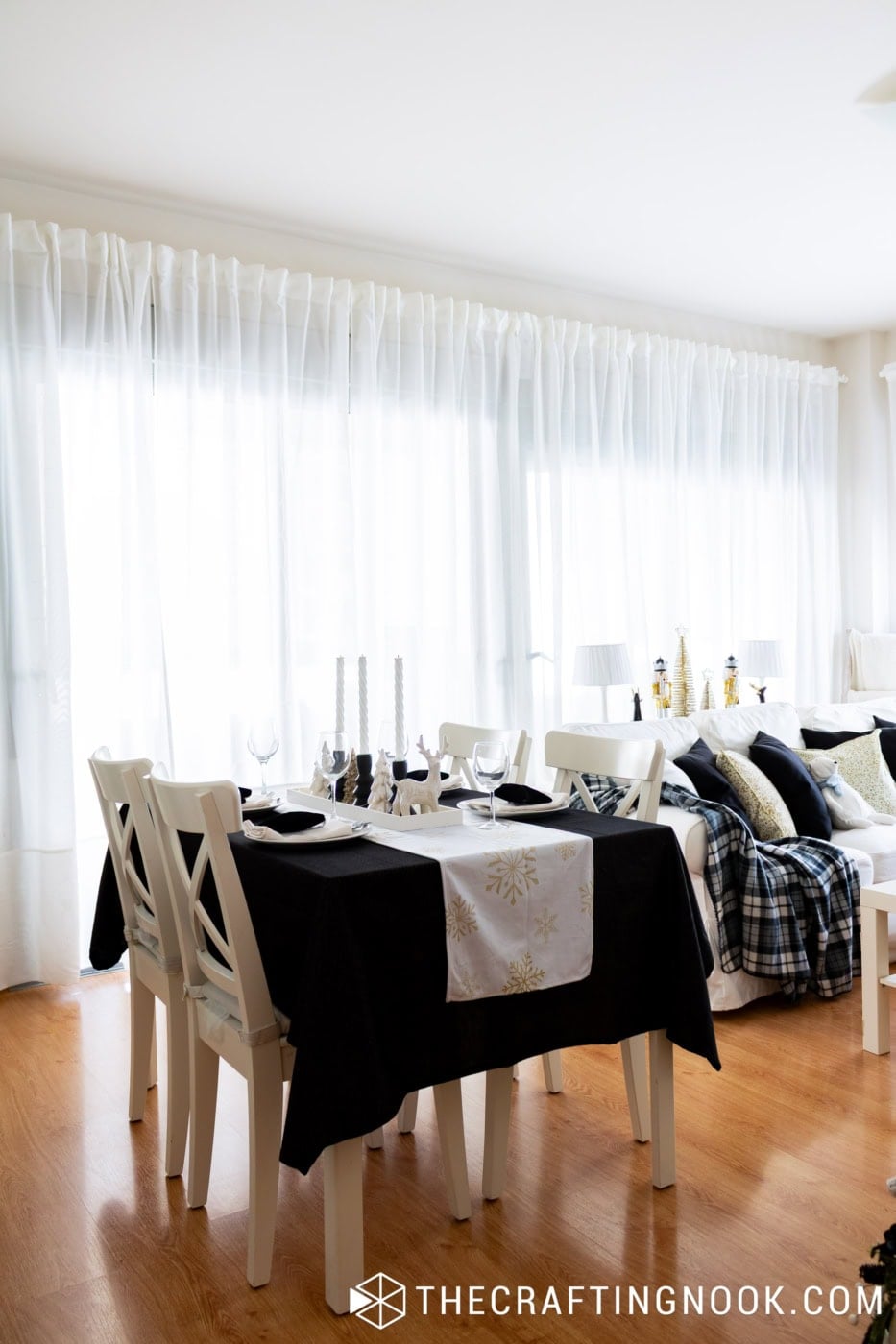 Elegant black and white Christmas dining table with white chairs, gold snowflake table runner, and Christmas decorations beside the sofa.
