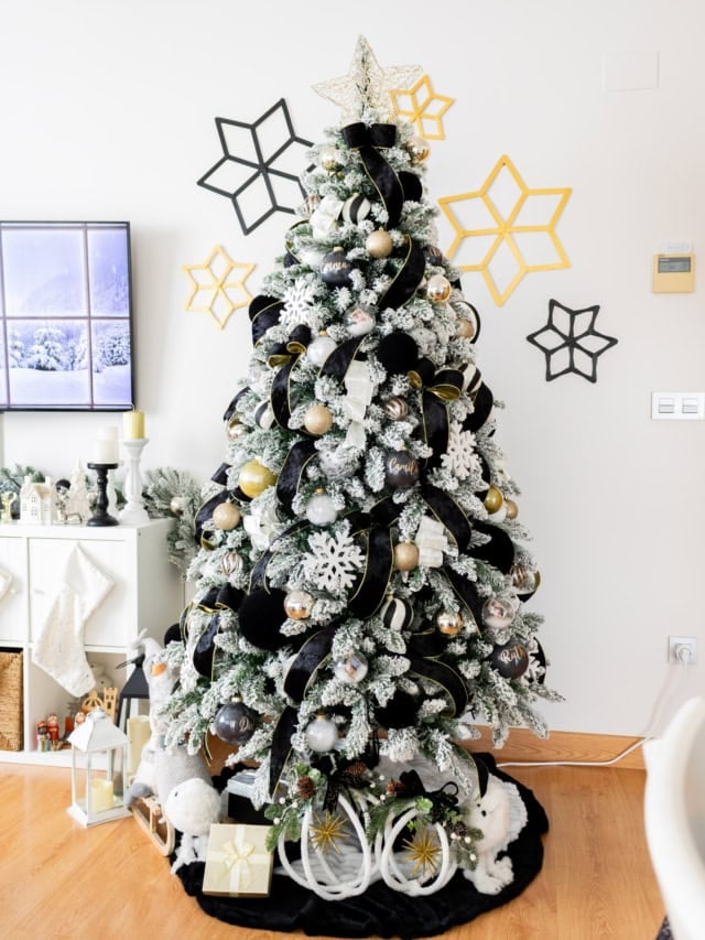 Elegant black, white and gold Christmas tree with velvet ribbons, gold ornaments and snowy flocked branches in a decorated living room.