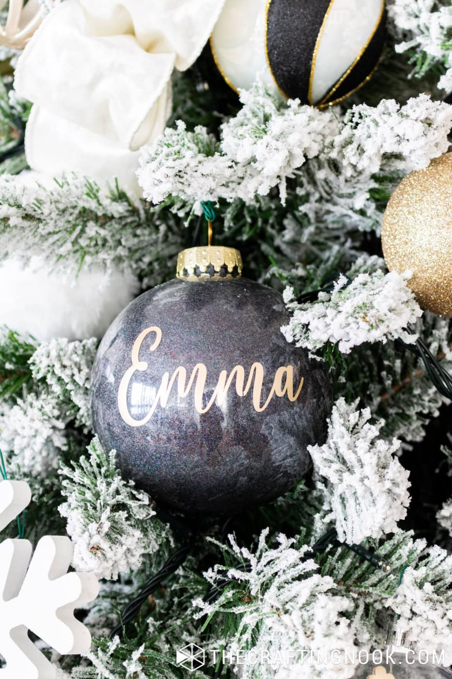 Black glitter Christmas ornament personalized with Emma in white vinyl lettering on a flocked Christmas tree.