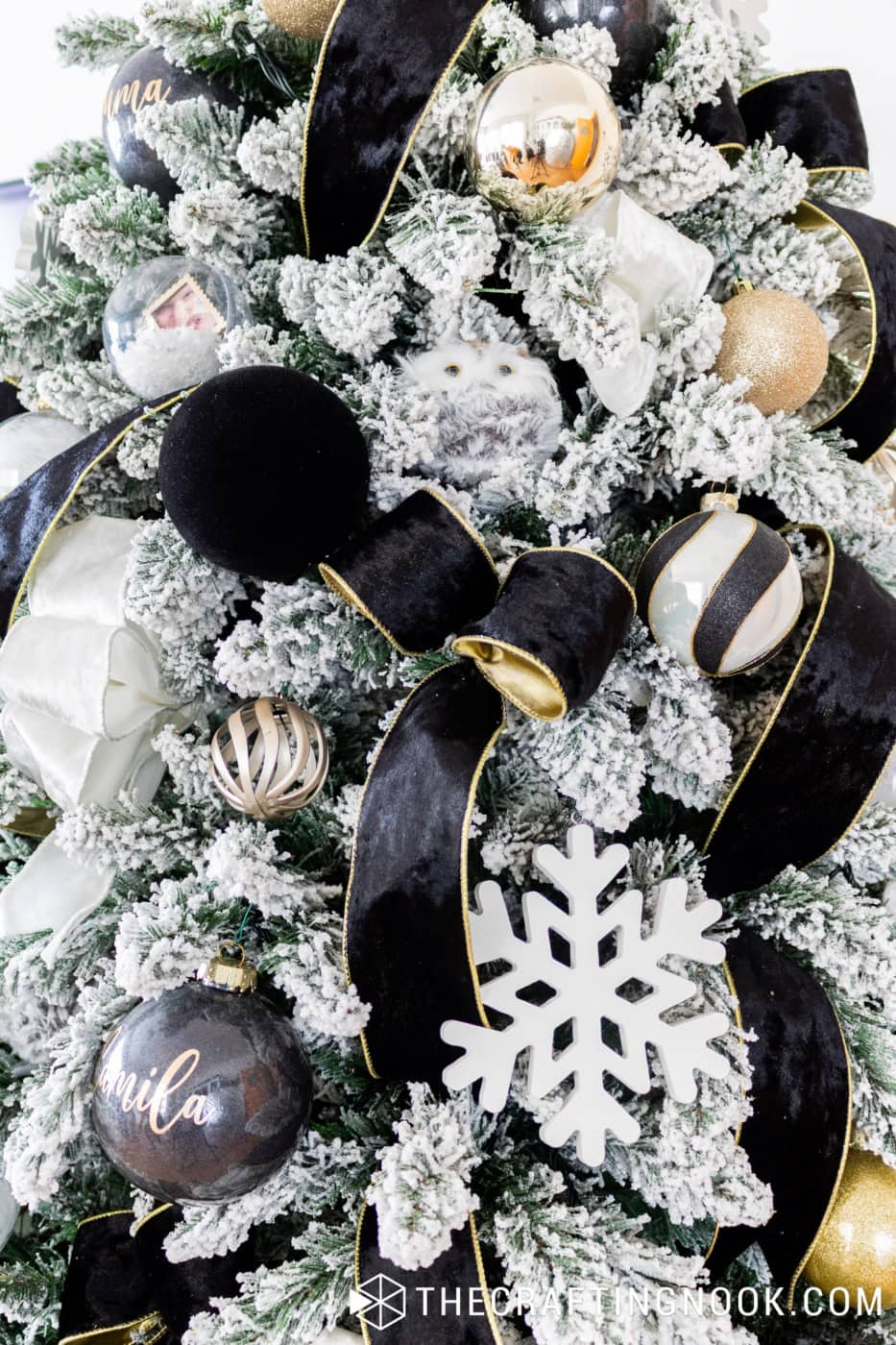 Wide black velvet ribbon with gold trim, white ribbon, black velvet ornaments and gold baubles on snowy Christmas tree branches.