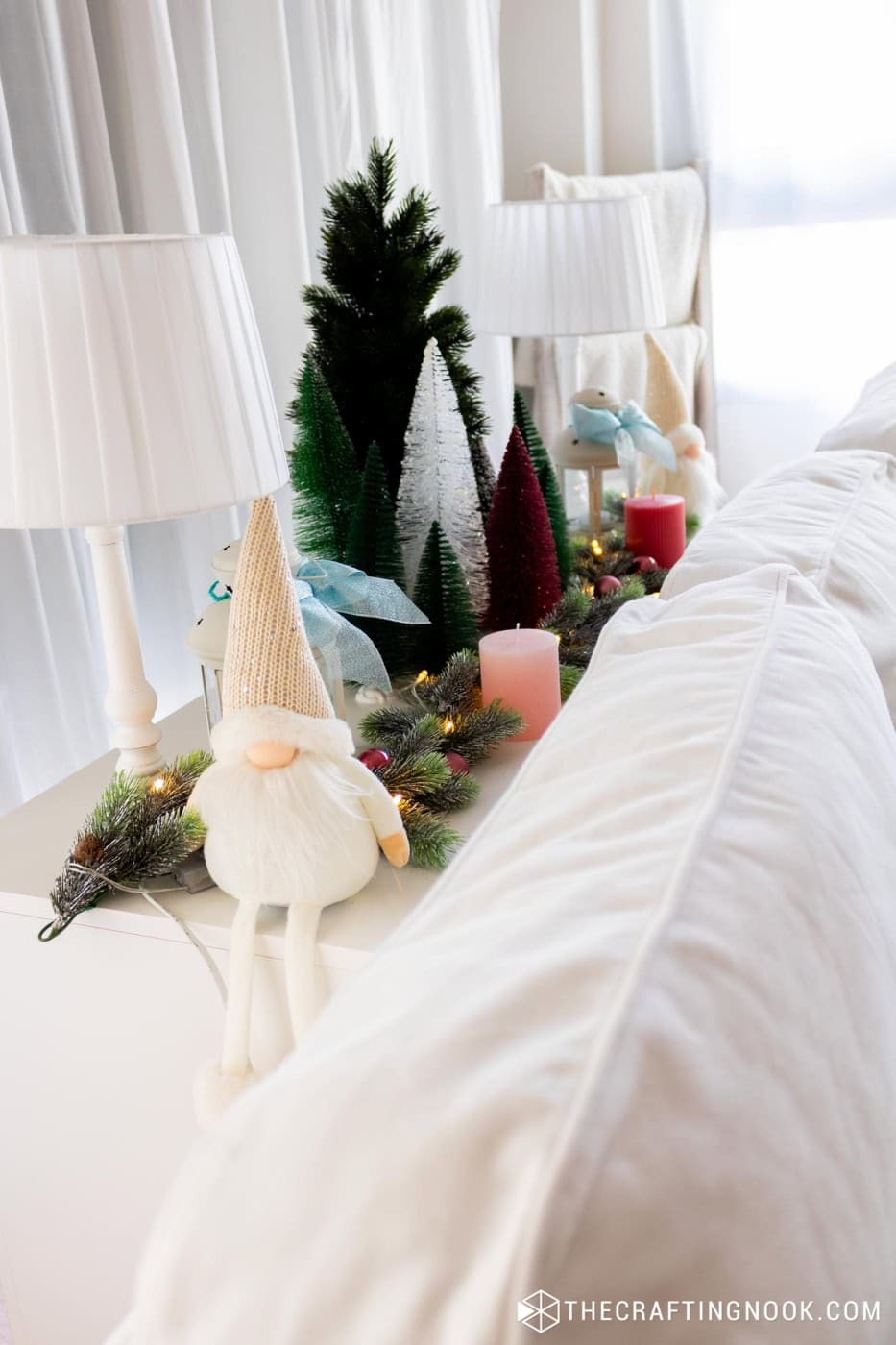 White gnome sitting next to green, white and burgundy bottle brush trees, candles and a small garland